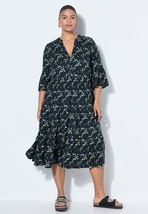 Frau trägt ein schwarzes Midikleid mit kleinem grünem und blauem Blumenmuster, ausgestellten Ärmeln und schwarzen Plateau-Sandalen.