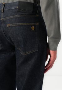 Dark blue denim jeans with orange stitching, featuring a black leather waistband tag, back pockets, and a small logo patch.