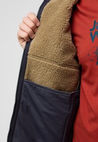 Veste zippée bleu marine avec une doublure en polaire texturée beige et une poche intérieure. La couche extérieure est lisse avec une finition mate.