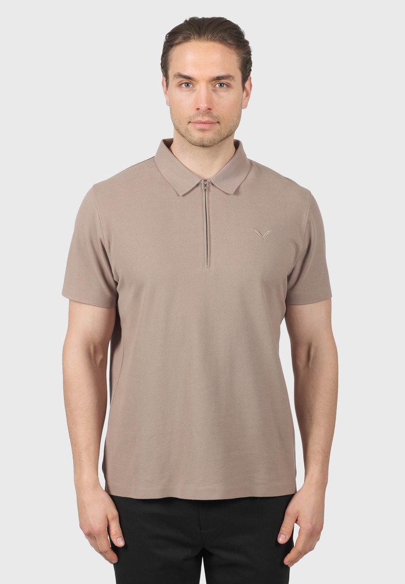 Homme aux cheveux bruns portant un polo beige à manches courtes avec une fermeture éclair à l'avant, debout devant un fond gris clair uni.