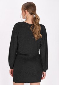 Svart klänning med långa, puffiga ärmar och markerad midja. Tyget har en subtil glans som ger en strukturerad look.