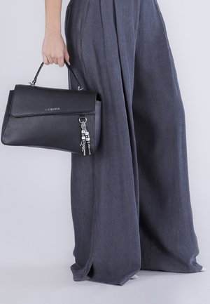 Persona con pantalones grises de pierna ancha sosteniendo un bolso de cuero negro con un llavero y herrajes plateados.