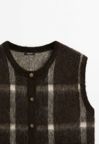 Veste sans manches marron foncé duveteuse avec motif à carreaux beige et gris et trois boutons décoratifs dorés sur le devant.