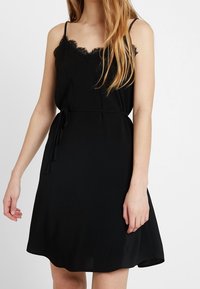 Schwarzes ärmelloses Kleid mit Spitze am Ausschnitt, Stoffgürtel in der Taille und einem fließenden A-Linien-Schnitt aus weichem Material.