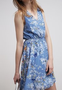 Hellblaues Blumen-Kleid mit V-Ausschnitt, elastischem Taillenbund und ausgestelltem Rock. Verfügt über weiße und bunte Blumenmuster auf der gesamten Fläche.