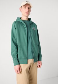 Zeleni zip-up dukserica napravljena od mekanog materijala s prednjim džepom, sa logotipom na prsima, preko odgovarajuće majice.