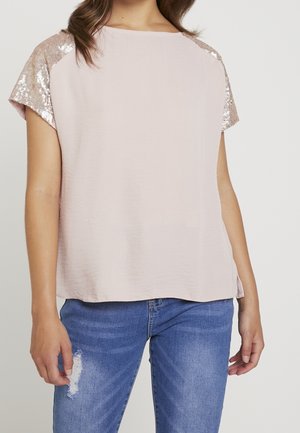 Nainen, joka pukeutunut vaaleanpunaiseen lyhythihaiseen toppiin, jossa on ruusukultaiset paljettiolkaimet, ja sinisiin kuluneisiin farkkuihin, seisoo yksinkertaisen taustan edessä.