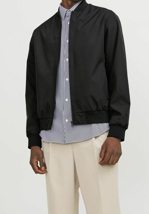 Homme portant une veste bomber noire sur une chemise à rayures noires et blanches boutonnée, avec un pantalon beige, sur un fond uni.