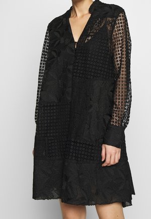 Schwarzes Spitzenkleid mit floralen und geometrischen Mustern, langen transparenten Ärmeln und Kragen, getragen von einer teilweise sichtbaren Person.