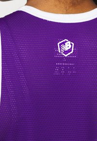 Fialový sportovní dres z prodyšné síťoviny s bílým lemováním. Na zádech bílá šesterečná značka. Texturovaná, prodyšná látka.