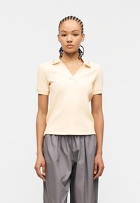 Γυναίκα με πλεξούδες, ντυμένη με ανοιχτό μπεζ κοντομάνικο ριμπ μπλουζάκι και άνετο γκρι παντελόνι, στέκεται μπροστά σε απλό φόντο.