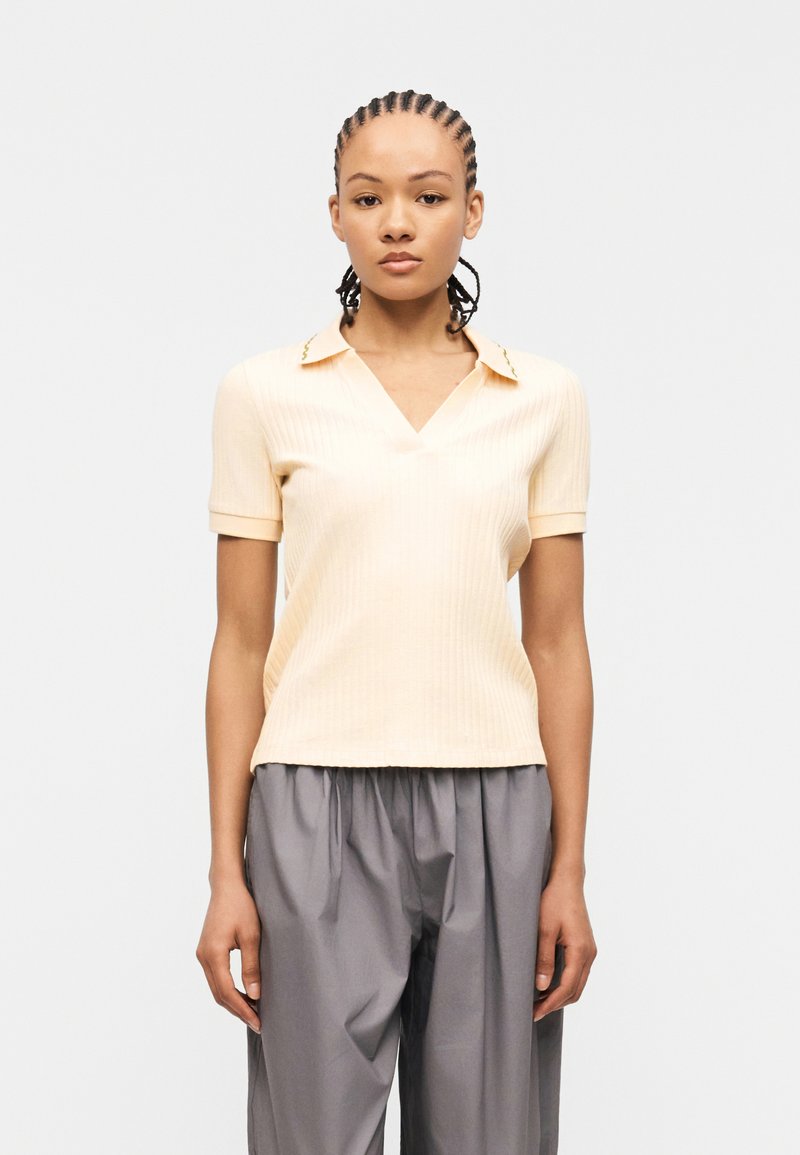 Γυναίκα με πλεξούδες, ντυμένη με ανοιχτό μπεζ κοντομάνικο ριμπ μπλουζάκι και άνετο γκρι παντελόνι, στέκεται μπροστά σε απλό φόντο.