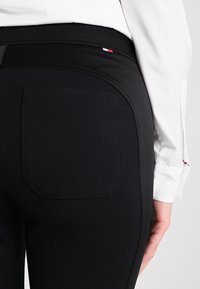 Close-up van een persoon in zwarte broek met een achterzak en een klein rood, wit en blauw vlaglogo nabij de tailleband, gecombineerd met een wit shirt.