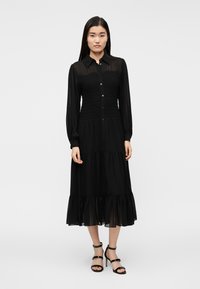 Čierne dlhé šaty s nariaseným živôtikom na gumičku, zapínaním na gombíky, stupňovitým lemom a priehľadnou látkou. Kombinované s čiernymi sandálmi na podpätku so strapcami.