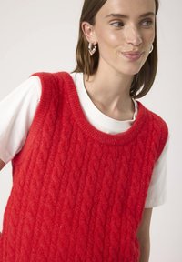 Woman wearing a bright red textured knit sleeveless sweater over a white short-sleeve shirt with silver hoop earrings.