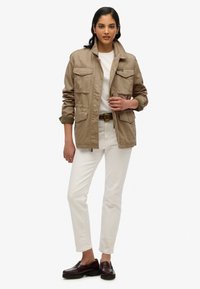 Beige utilityjack met meerdere voorkanten zakken, gecombineerd met een wit shirt en crèmekleurige broek. Bruine instappers maken de outfit compleet.