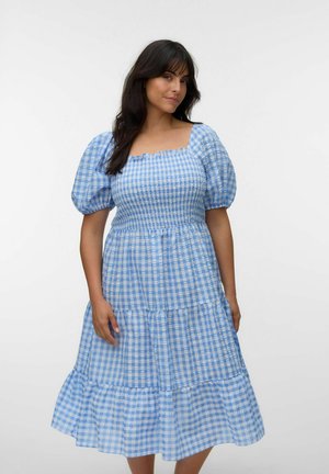 Kvinde iført en blå og hvid ternet midikjole med pufærmer og en rynket overdel mod en ensfarvet hvid baggrund.