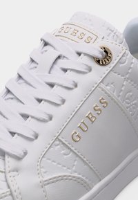 Biele tenisky Guess s reliéfnym vzorom, zlatými písmenami loga na boku a jazyku, bielymi šnúrkami a zlatým detailom na očku.