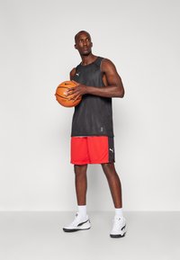 Svart sports tank topp med grå detaljer, røde treningsshorts og hvite joggesko. Holder en oransje basketball. Flat bakgrunn.