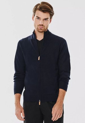 Granatowy sweter zapinany na zamek z wysokim kołnierzem, prążkowanymi mankietami i dołem. Wykonany z miękkiego materiału, z detalem w postaci skórzanego uchwytu zamka.