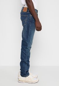 Homme portant un jean bleu en denim et des baskets blanches, debout de côté devant un fond blanc uni.