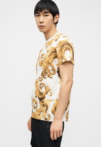 Νέος άνδρας με ίσια μαύρα μαλλιά που φοράει λευκό μπλουζάκι με χρυσά και καφέ περίτεχνα σπειροειδή μοτίβα και μαύρο παντελόνι, με φόντο απλό.