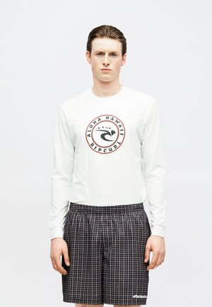 Νέος άνδρας που φοράει λευκό πουκάμισο με μακριά μανίκια με το λογότυπο "Aloha Hawaii Rip Curl" και μαύρο καρό σορτς, στέκεται μπροστά σε απλό φόντο.