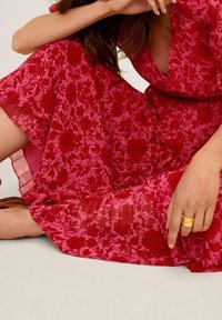 Robe fleurie en rose vif avec des motifs de fleurs rouges, manches courtes et une coupe fluide. Le matériau semble léger et transparent. Une bague en or est visible.
