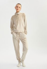 Beige atletisk sæt bestående af en højhalset pullover med halvlukning og matchende joggers med lommer, lavet af blødt, tekstureret stof.