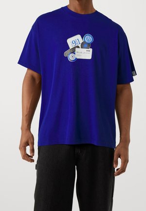 Male wearing a royal blue t-shirt with cluster of graphic patches including “BLUE JEANS” text, paired with black pants, shown from shoulders to hips.