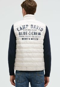 Mężczyzna ubrany w biały pikowany kamizelkę z napisem "Camp David Sailing Club Blue Crew" i logo kotwicy z tyłu, granatowymi rękawami oraz niebieskimi dżinsami.