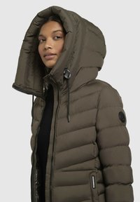 Olive green puffer jacket with a large hood, quilted design, zip closure, and drawstring. Features a logo patch on the sleeve.