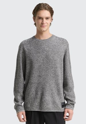 Junger Mann mit kurzen braunen Haaren, der einen langärmeligen grauen Strickpullover und schwarze Hose trägt, steht vor einem einfarbigen hellen Hintergrund.