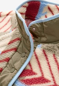 Col et partie supérieure d'une veste polaire avec tissu matelassé vert olive et motif géométrique rouge, crème et bleu, fermé par des boutons-pression.