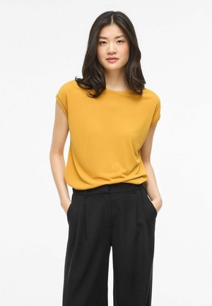 Jauna moteris su ilgais tamsiais plaukais, dėvinti garstyčių geltonumo trumpomis rankovėmis palaidinę ir juodus aukštos juosmens kelnes, rankos kišenėse.