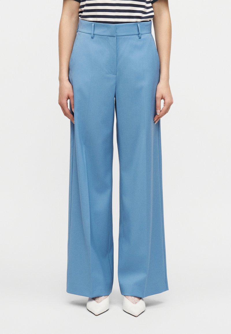 Γυναίκα που φοράει ψηλόμεσα, φαρδι ά παντελόνια σε ανοιχτό μπλε, ριγέ μπλούζα και λευκά μυτερά παπούτσια με δαντελένια λεπτομέρεια, στέκεται μπροστά σε λευκό φόντο.