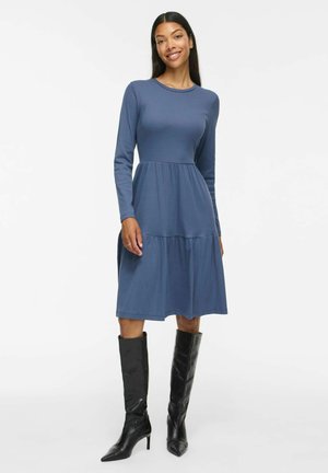 Femme portant une robe bleue à manches longues, longueur genou, et des bottes noires à talons montant jusqu'aux genoux, debout et souriante sur fond blanc.