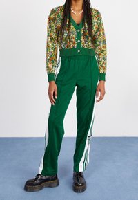 Vihreä harjoituspuku kukkakuosisella neuletakilla, jossa on napit. Housuissa on valkoisia raitoja. Mallilla on päällään tukevat mustat kengät.