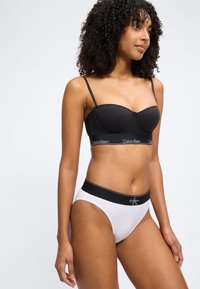 Γυναίκα που φοράει μαύρο σουτιέν Calvin Klein και λευκά ψηλόμεσα εσώρουχα με μαύρη μέση, στέκεται μπροστά σε απλό ανοιχτό φόντο.