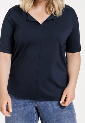 Vrouw die een donker marineblauw kortemouw V-hals topje en blauwe spijkerbroek draagt, getoond van schouders tot heupen tegen een effen achtergrond.