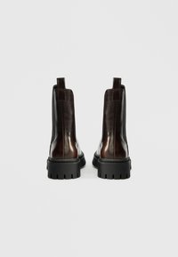 Καφέ δερμάτινες μπότες μέχρι τον αστράγαλο με γυαλιστερή επιφάνεια, εφοδιασμένες με χοντρή μαύρη καουτσούκ σόλα, ελαστικά πλαϊνά τμήματα και θηλιές από πίσω.