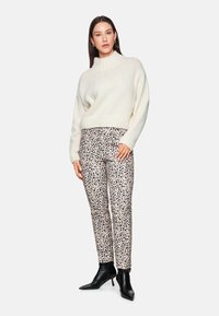 Cremfarbener Strickpullover mit hohem Kragen, kombiniert mit beigen Hosen mit schwarzem Tierdruck. Schwarze Stiefeletten runden das Outfit ab.