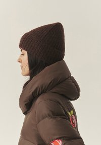 Barna kötött sapka hegyes tetejével, sötétbarna pufi dzsekivel viselve, amelynek ujján színes virágos hímzés látható.