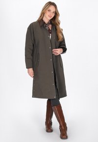Olijfgroen lange jas met een knoopsluiting aan de voorkant, bruine kraag en zijzakken, gedragen over grijze leggings en bruine kniehoge laarzen.