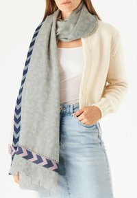 Hellgrau Schal mit strukturiertem Muster und marineblauem Chevron-Design, ergänzt mit rosa und grünen Akzenten, getragen mit einem cremefarbenen Cardigan und Jeans.