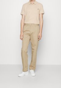 Beige polo tričko s límečkem a kapsou; hnědé chino kalhoty; bílé tenisky. Hladká tkanina a přiléhavý střih s jemným detaily na tričku.