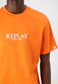 Oranžinė medvilninė marškinė su baltais užrašais "REPLAY" ir "SINCE 1981". Trumpomis rankovėmis, apvaliu kaklu, ant rankovės yra mažas prekės ženklo etiketė.