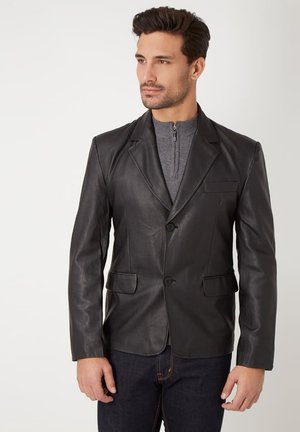 Hombre que lleva una blazer de cuero negra sobre un jersey gris con cremallera en el cuello y vaqueros oscuros, mirando ligeramente hacia la derecha sobre un fondo liso.