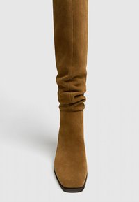 Botte haute en daim marron montant jusqu'au genou, avec un bout carré arrondi et un design froncé. Texture lisse et semelle plate avec des éléments métalliques minimalistes.