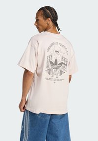Ljusrosa oversized t-shirt med en grafisk design på baksidan med texten "ORIGINALS FESTIVAL", tillsammans med stadnamn och konstverk.